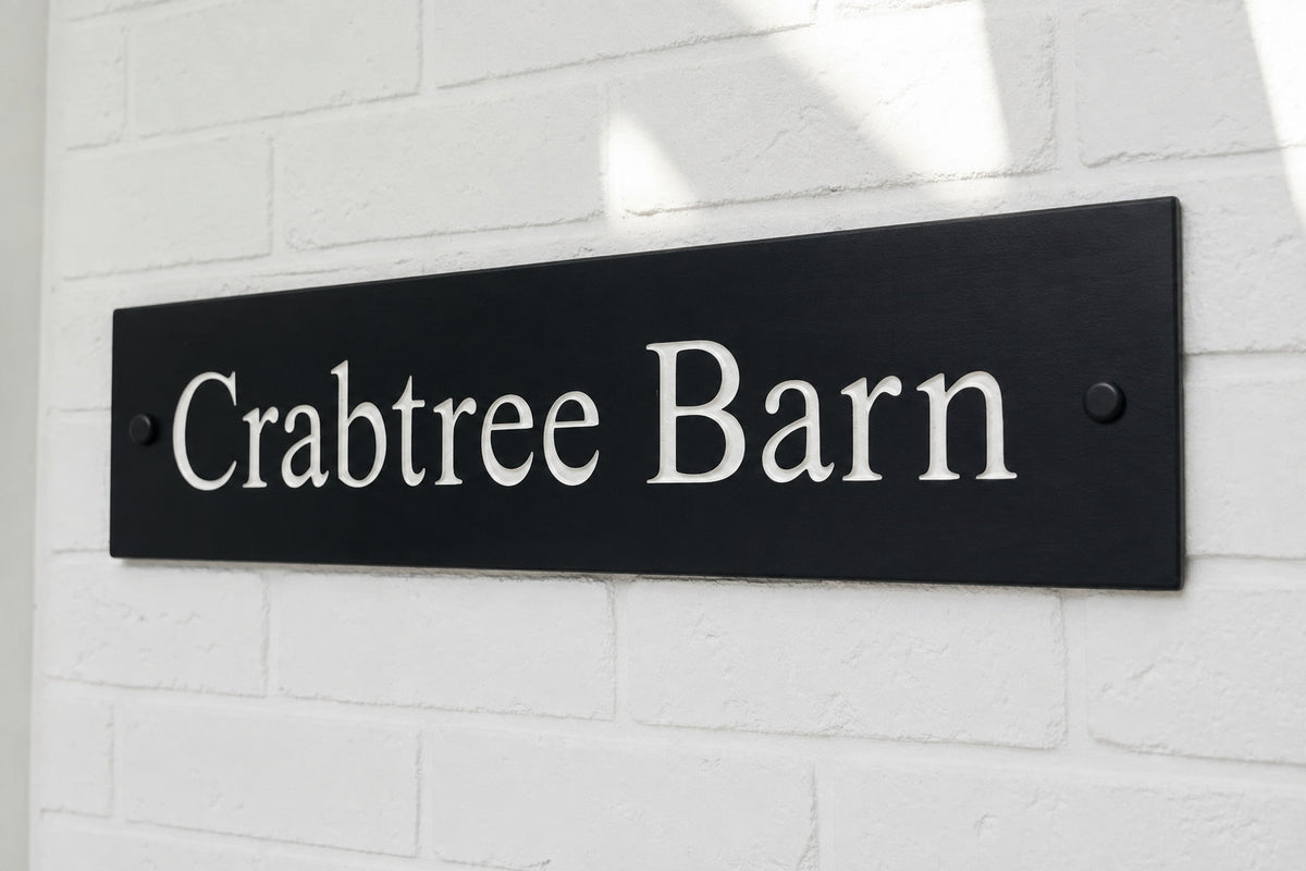 Natural slate house name sign reading ‘Crabtree Barn’, with pale lettering and black fixing caps on white brickwork.