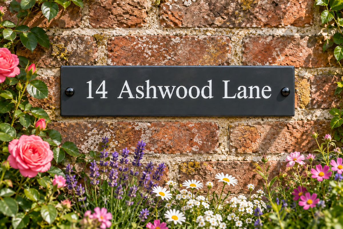 Natural slate address sign reading ‘14 Ashwood Lane’, mounted on a brick garden wall surrounded by flowers.
