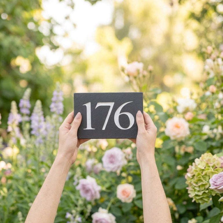 Hands holding a natural slate house number 176 sign in a flower garden.